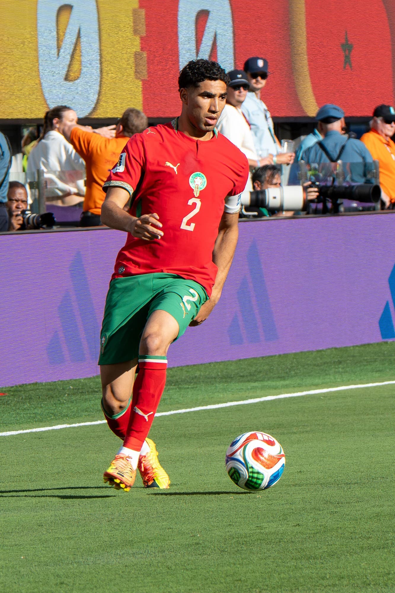 Achraf Hakimi me fanellën e kuqe të Marokut pranë topit në fushën e futbollit.