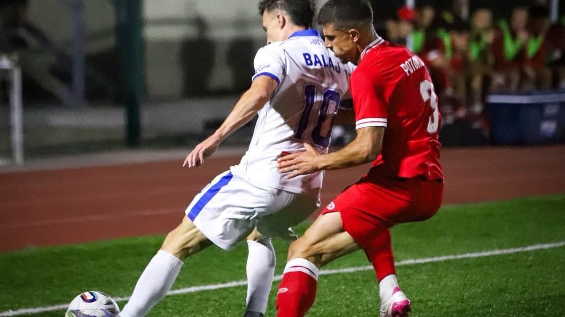 Dy futbollistë, njëri me uniformë të bardhë e blu dhe tjetri me të kuqe, në duel për topin në fushë.