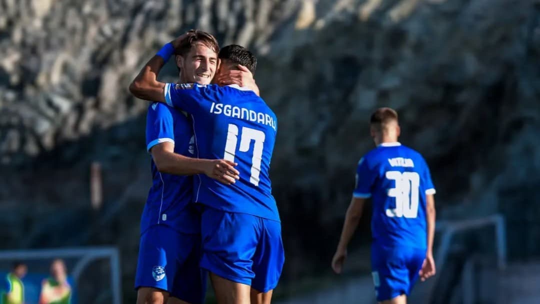 Dy futbollistë me fanella blu përqafohen në fushë, ndërsa një bashkëlojtar shihet në sfond.