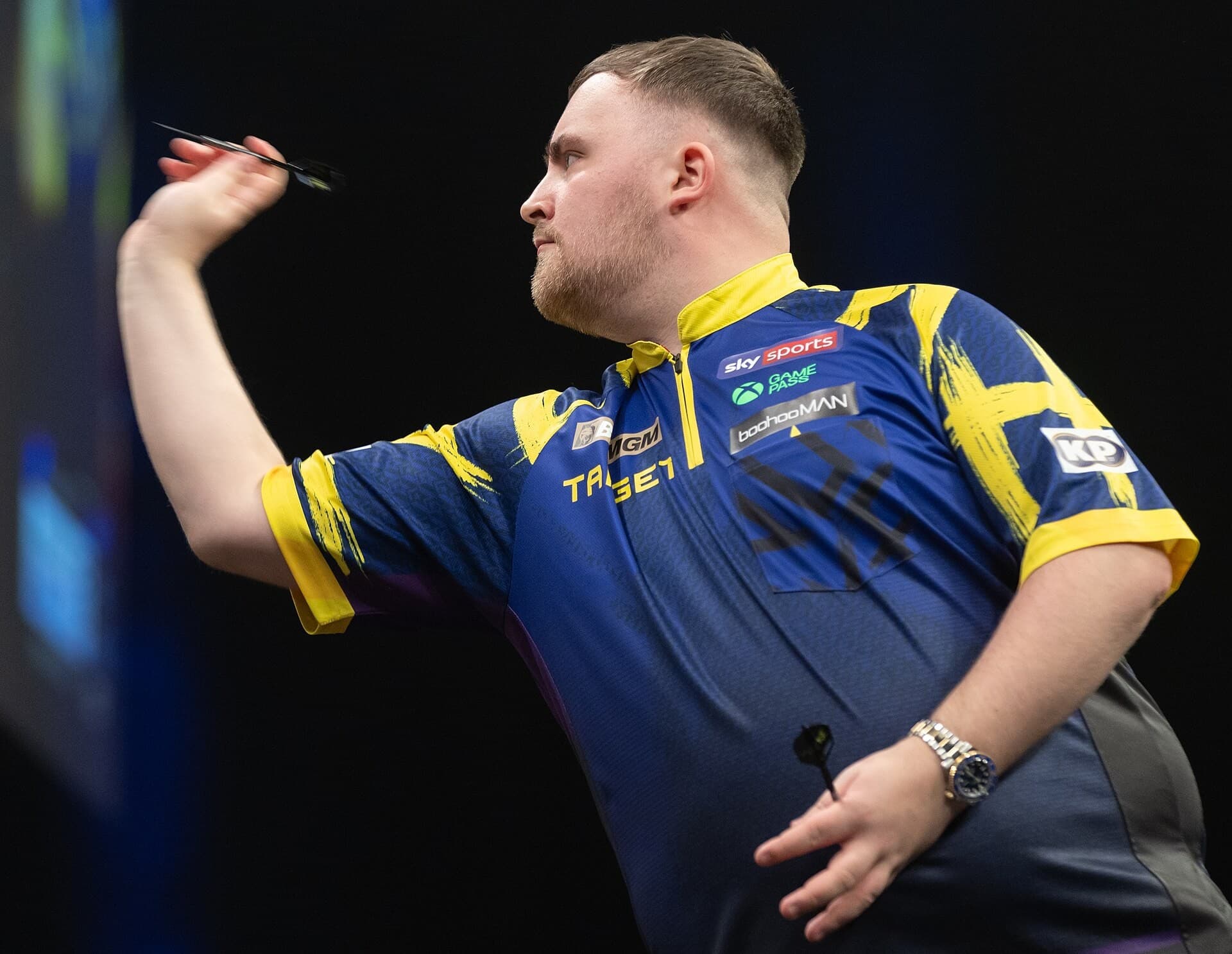 Luke Littler me fanellë blu dhe të verdhë, duke hedhur një shigjetë gjatë Premier League Darts në Berlin, më 26 mars 2026.