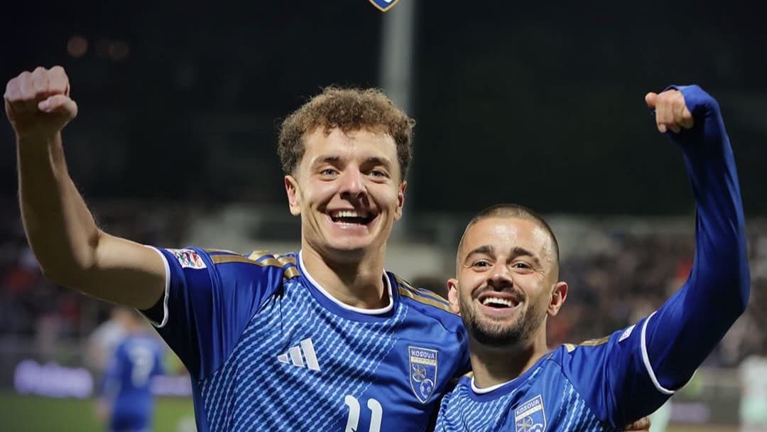 Dy futbollistë me fanellat e Kosovës, të buzëqeshur dhe me krahët e ngritur në stadium.
