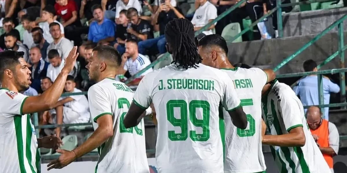 Futbollistë me fanella të bardha e të gjelbra të mbledhur pranë tribunës me spektatorë.