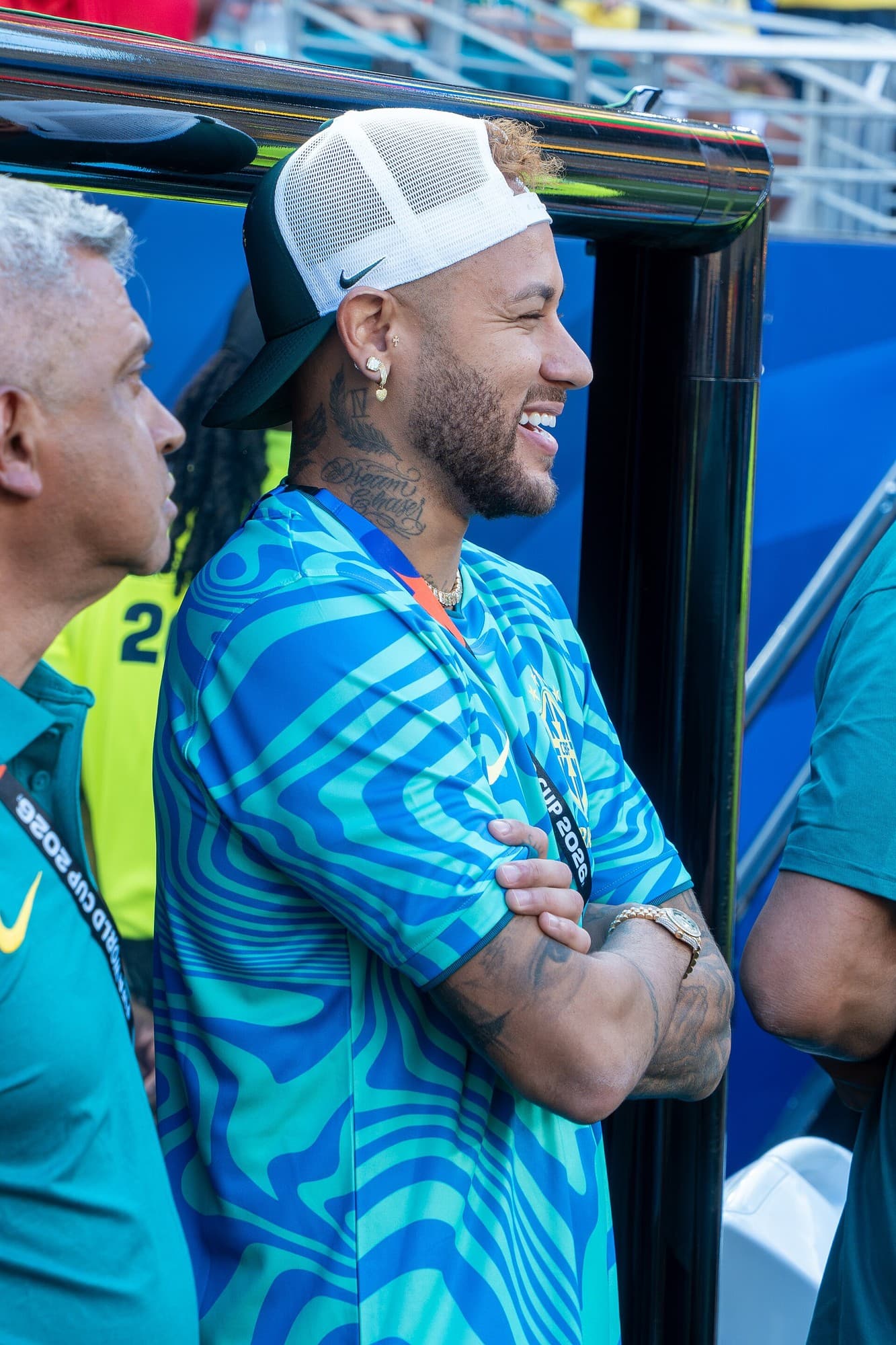 Neymari me veshje sportive të Brazilit dhe kapele të kthyer mbrapsht, me krahët e kryqëzuar pranë stolit.