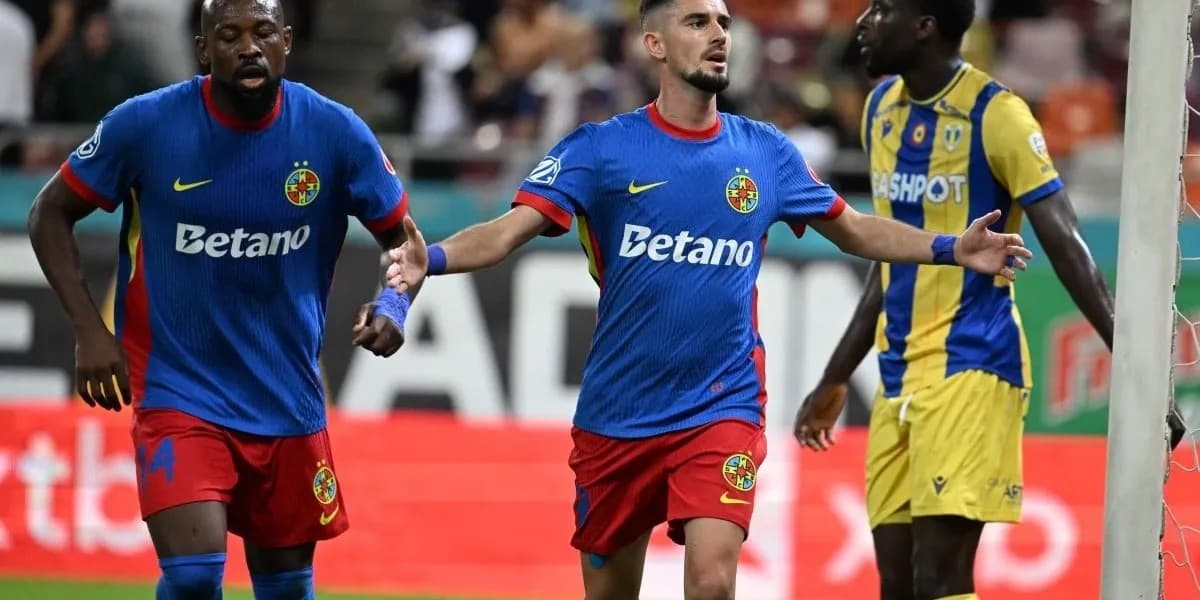 Futbollisti në qendër me fanellë blu dhe pantallona të kuqe mban krahët hapur, pranë dy lojtarëve të tjerë dhe shtyllës së portës.